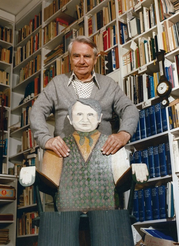 Zu sehen ist Rolf Italiaander im älteren Alter zuhause vor seiner Bibliothek stehend. Mit weißem, vollem Haar, einem weißen, gepunkteten Hemd und einer hellgrauen Strickjacke an. Vor ihm steht ein angemalter Stuhl aus Holzplatten bestehend, der ihn selbst sitzend darstellt. Aus der breiten Lehne ragt sein gemalter Kopf hervor, die Armlehnen stellen seine Arme dar. Rolf Italiaander greift von hinten auf die Lehne des kreativen Stuhls.