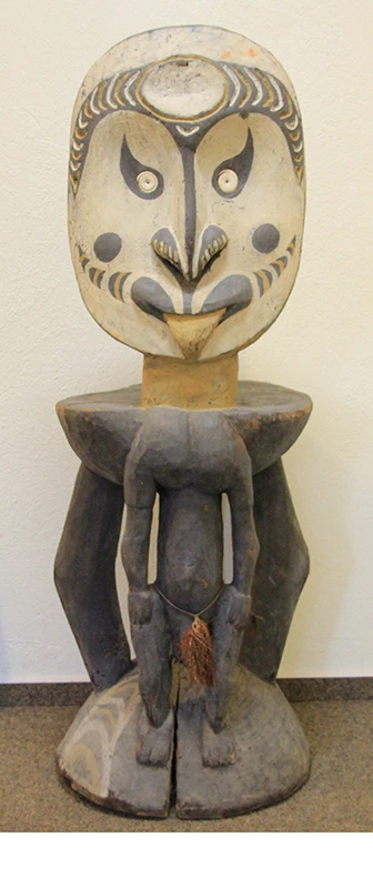 Kulthocker aus dem Sepik-Gebiet in Papua-Neuguinea: Ein dunkler Holzhocker mit drei Beinen und einem großen ovalen Gesicht als Lehne. Das Gesicht ist weiß bemalt mit feinen dunklen Zügen und streckt die Zunge heraus. Das vordere Bein des Hockers stellt einen kleinen Körper dar, dessen fehlender Kopf durch das Gesicht der Lehne ergänzt wird.