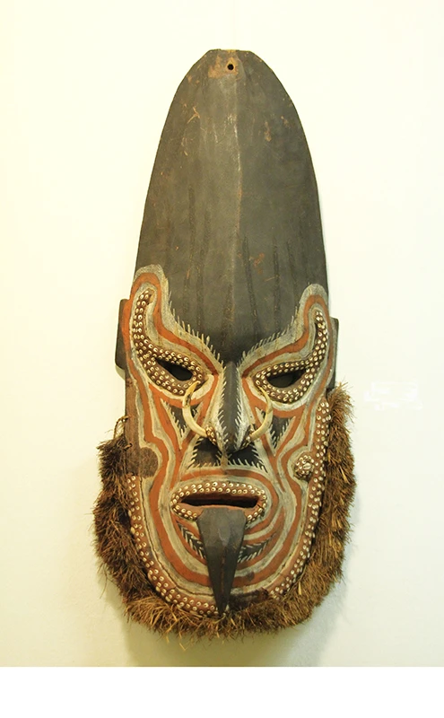 Holzschild: Ein lang gezogenes Gesicht mit einer hohen, dunklen Stirn. Das Gesicht ist mit roten Linien bemalt und mit kleinen Muscheln verziert, der Bart ist aus Stroh. Aus dem Mund wird die Zunge ausgestreckt.