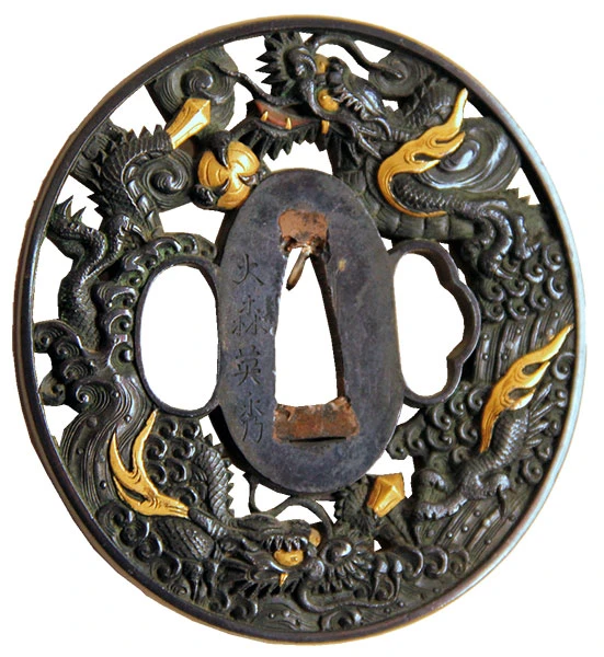Ein verziertes Schwertstichblatt der traditionellen japanischen Schwerter, genannt Tsuba, meist aus Metallen wie Eisen, Kupfer oder Kupferlegierungen. Die schwarze ovale durchbrochene Scheibe besteht aus verschlungenen Drachen, die vergoldetes Feuer speien. In der Mitte der Scheibe sind drei Öffnungen für die Klinge. Sie dient als Schutz zwischen Klinge und Griff.
