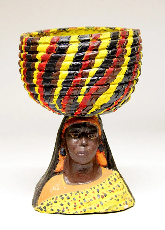Büste einer afrikanischen Frau, die auf ihrem Kopf einen riesigen, geflochtenen Korb trägt. Der Korb ist mit gelben, schwarzen, roten und braunen Streifen bemalt. Sie hat ein Tuch um ihren Kopf und ein gepunktetes Tuch um ihre Schultern. Das Gesicht ist fein und realistisch ausgearbeitet.