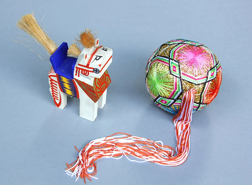 Zwei bunte Spielzeuge: Ein aus Holz geschnitztes weißes Pferd mit roten Verzierungen und einem blauen Sattel. Die Mähne und der Schweif sind aus Bast. Daneben ein Ball mit bunten Fäden in Mustern überzogen. Ein Band aus weißen und roten Fäden tritt aus dem Ball heraus.