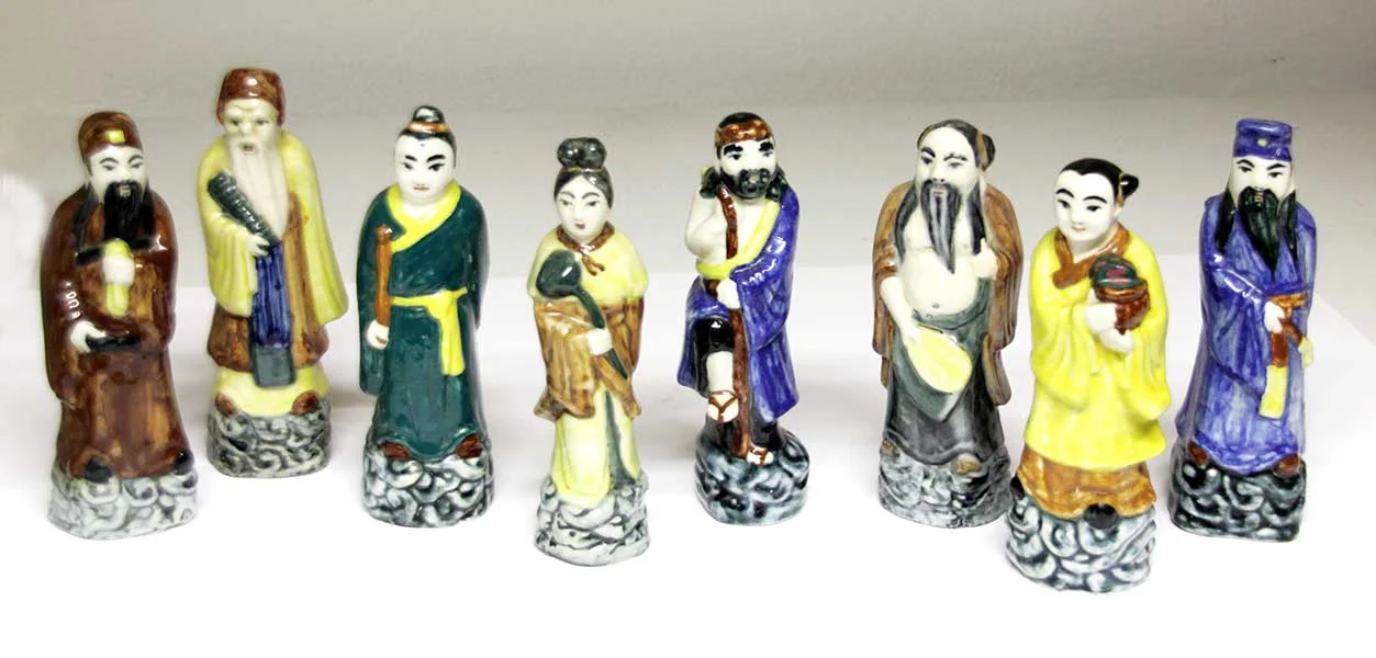 Acht chinesische kleine Figuren, die unterschiedlich gekleidete Männer mit Bärten und Frauen darstellen. Sie sind gebrannt und bemalt und stehen auf einem Sockel mit Wellenstruktur.