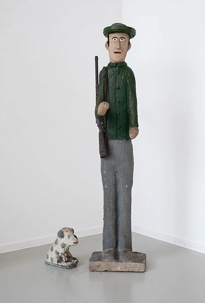 Eine schmale, hohe und simpel geschnitzte Holzfigur zeigt einen Jäger mit dunkelgrüner Jacke, Hut und einer grauen Hose auf einem flachen Sockel stehend. Über die Schulter hängt sein Gewehr herunter. Er guckt mit weißen runden Augen regungslos nach vorn. Neben ihm sitzt ein sehr keiner weißer Hund mit grauen Flecken.
